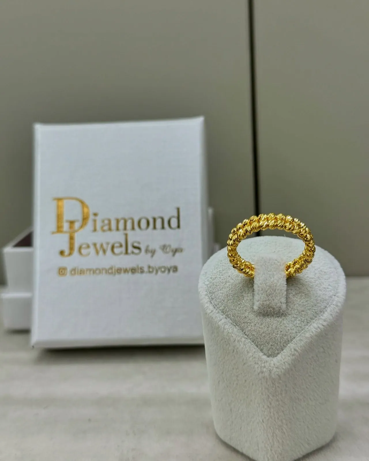 Geflochtener Goldring auf einem Ringständer vor einer Diamond-Jewels-Schachtel